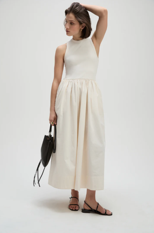 Woman wearing a white sleeveless top and beige skirt holding a black handbag on a white background