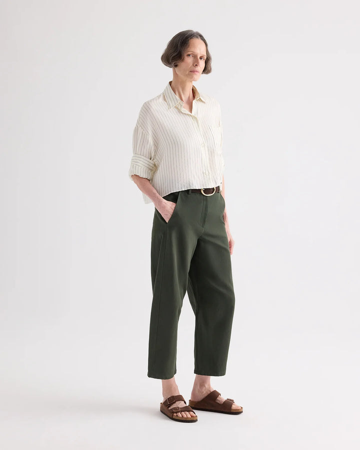 A woman stands in a studio wearing a cream striped button-up shirt, dark olive green cropped trousers, a thin belt, and brown slide sandals against a light gray background.