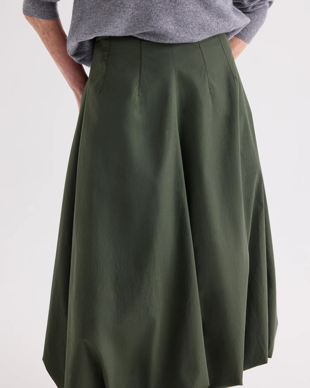 An olive green A-line skirt with front pleats, shown on a model wearing a gray long-sleeve top against a plain light gray background.