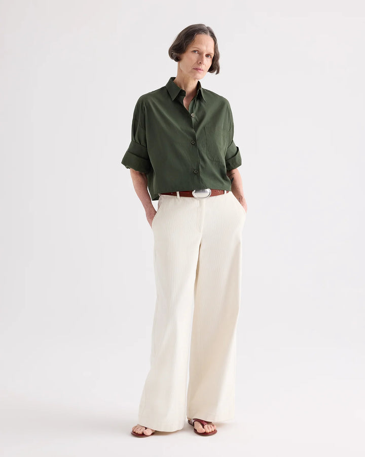 A woman modeling a dark olive green button-up shirt with rolled sleeves, paired with wide-leg cream trousers and brown sandals, against a plain white studio background.