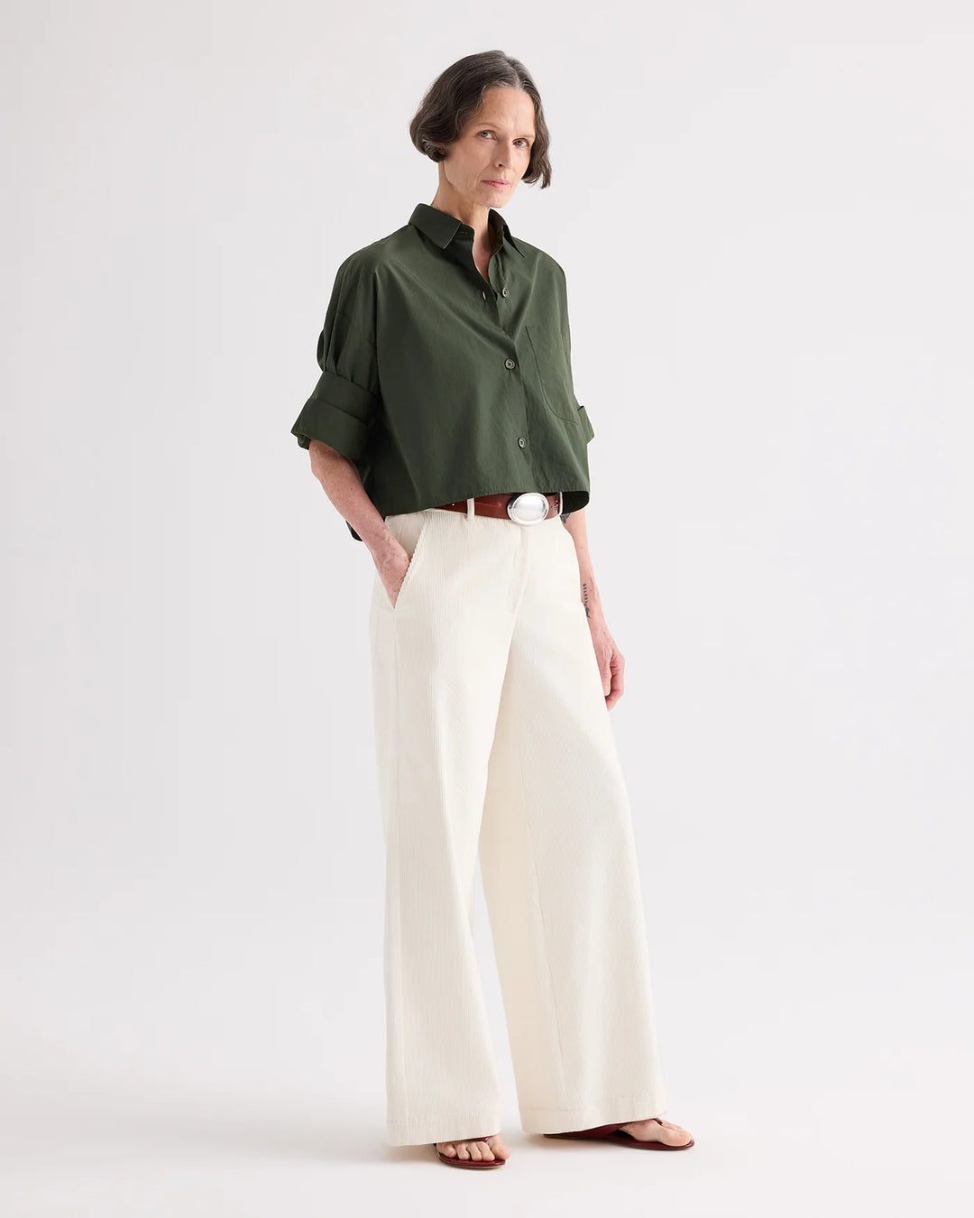 A model stands against a plain light gray studio background wearing a dark olive green short-sleeve button-up shirt, off-white wide-leg trousers, a brown belt, and flat sandals.