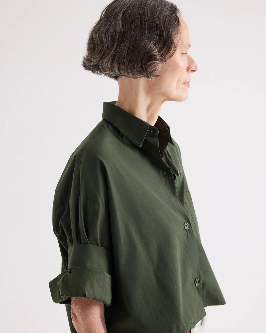 A woman in profile wearing an oversized dark olive green button-up shirt with rolled sleeves, photographed against a plain light gray studio background.