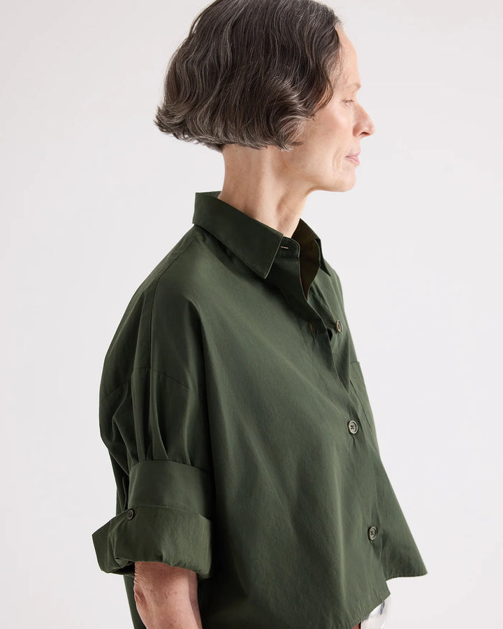 A woman in profile wearing an oversized dark olive green button-up shirt with rolled sleeves, photographed against a plain light gray studio background.