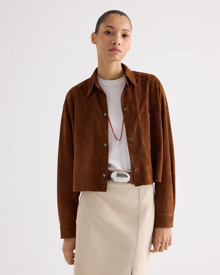 A model wearing a cropped brown suede button-up shirt jacket over a white t-shirt, paired with beige trousers and a silver belt, against a plain light gray background.