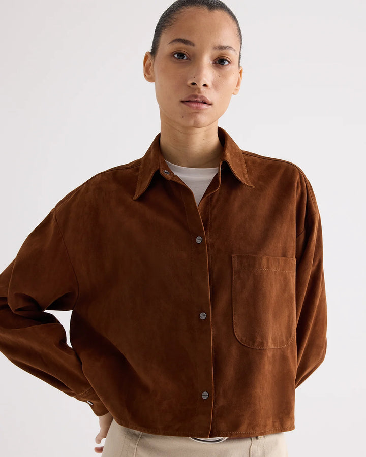 A woman wearing a brown suede button-up shirt over a white t-shirt and beige trousers, photographed against a light gray studio background.