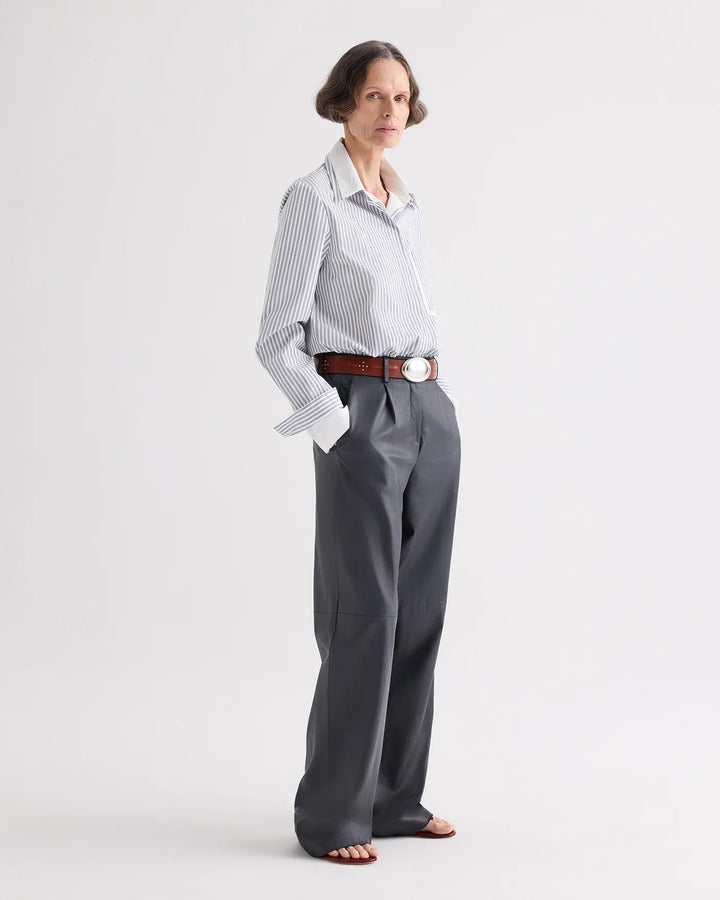 A woman modeling a light blue and white vertically striped button-down shirt tucked into high-waisted, wide-leg gray trousers, styled with a brown leather belt and brown sandals against a plain light gray studio background.