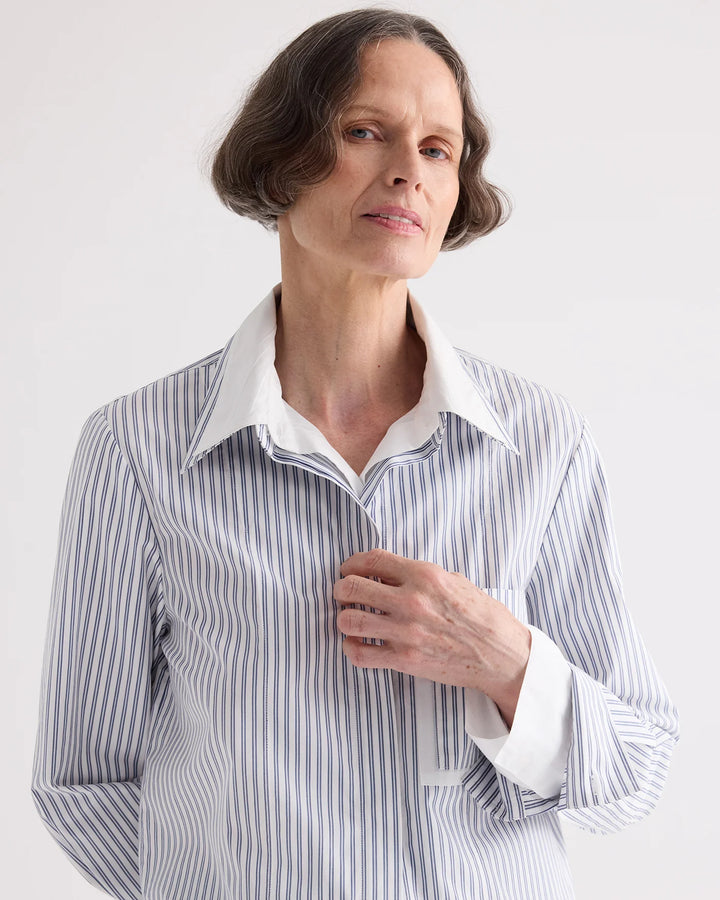 A woman wearing a long-sleeve button-up shirt with vertical blue and white stripes, featuring a solid white collar and cuffs, photographed against a light gray studio background.