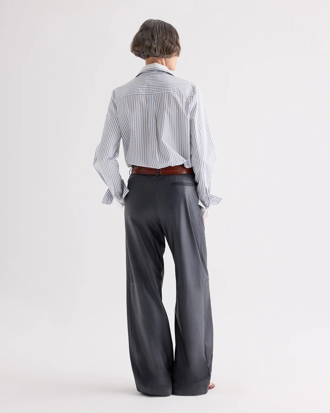 A model shown from the back wearing a light blue and white vertically striped button-down shirt, tucked into dark charcoal gray wide-leg trousers with a brown belt.
