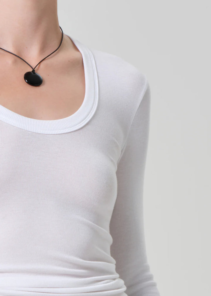 Person wearing a white long-sleeve shirt with a black necklace against a plain background