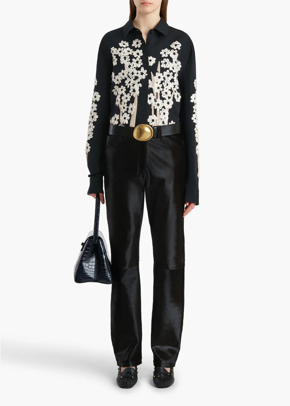 Person wearing a black floral blouse with white flowers, black pants, and holding a black handbag on a white background.