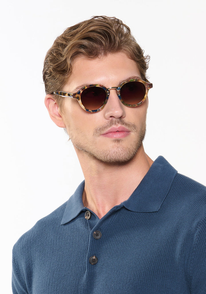 Man wearing tortoiseshell sunglasses and a blue polo shirt against a white background