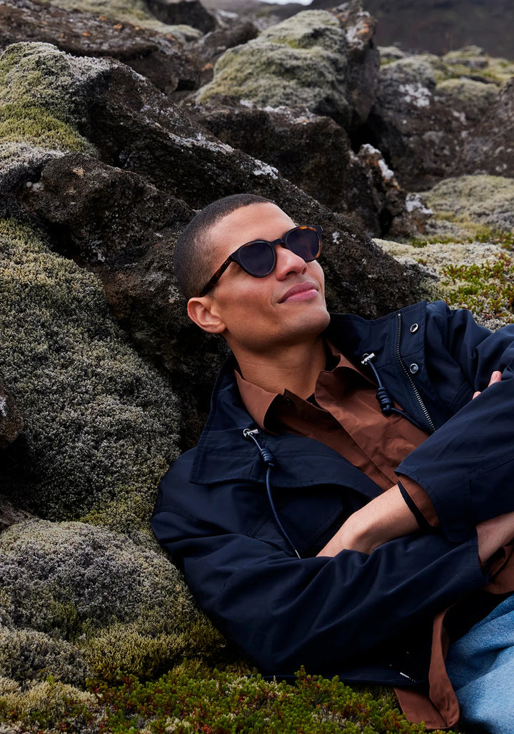 Man wearing sunglasses and a dark jacket sitting among moss-covered rocks.