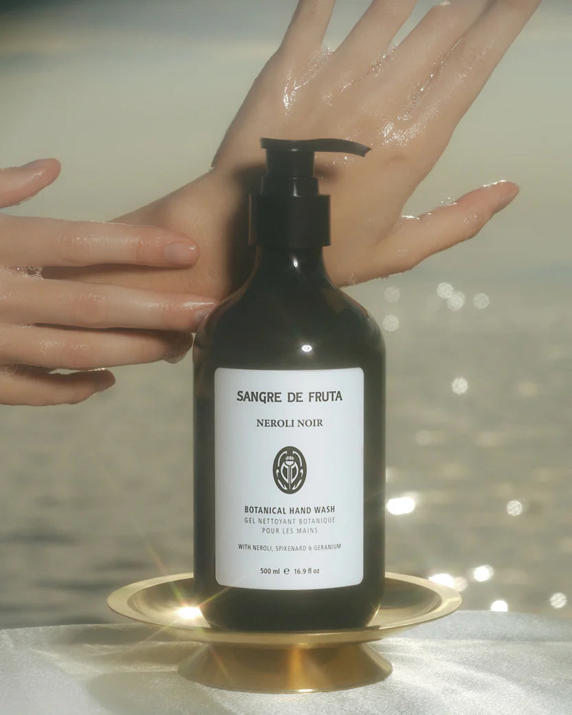 Bottle of Sangre de Fruta Neroli Noir hand wash on a reflective surface with hands washing them.