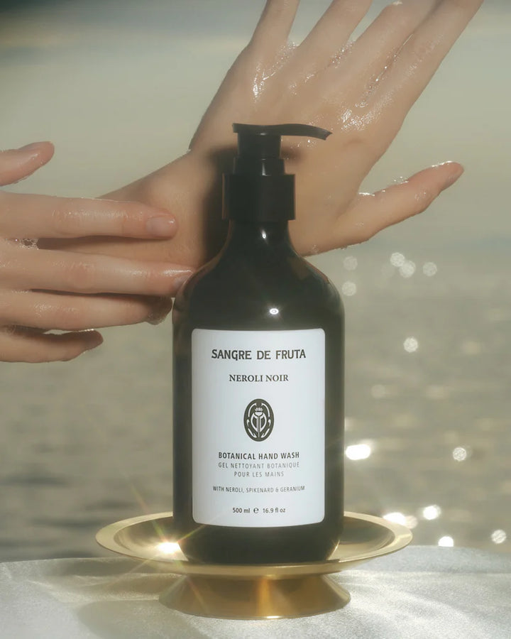 Bottle of Sangre de Fruta Neroli Noir hand wash on a reflective surface with hands washing them.