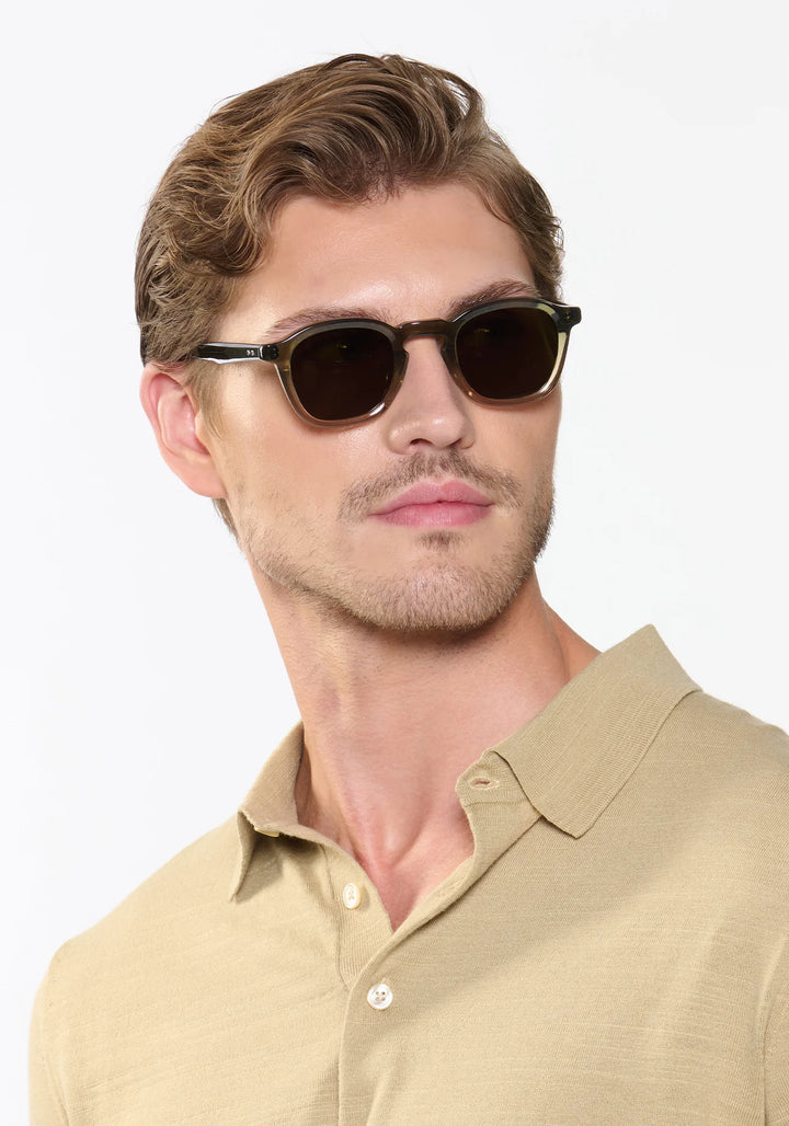 Man wearing dark sunglasses and a beige polo shirt against a light background