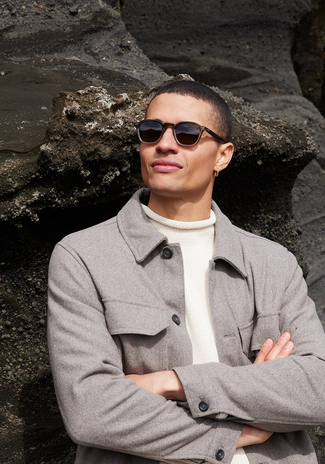 Man wearing sunglasses and a gray coat standing against a rocky background