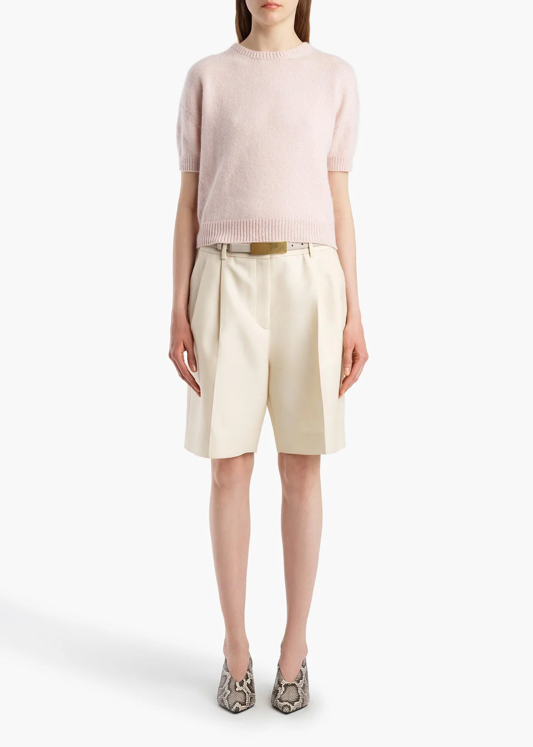 A model wearing a pale pink short-sleeve knit sweater, cream pleated shorts with a gold belt, and snakeskin-patterned heels, shot against a plain white studio background.