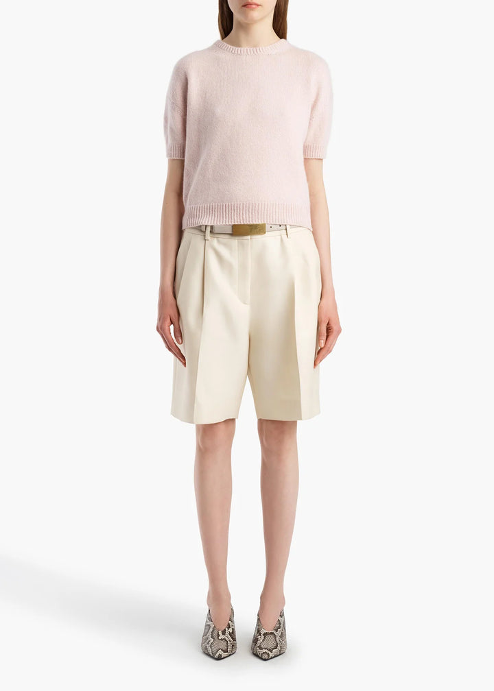 A model wearing a pale pink short-sleeve knit sweater, cream pleated shorts with a gold belt, and snakeskin-patterned heels, shot against a plain white studio background.