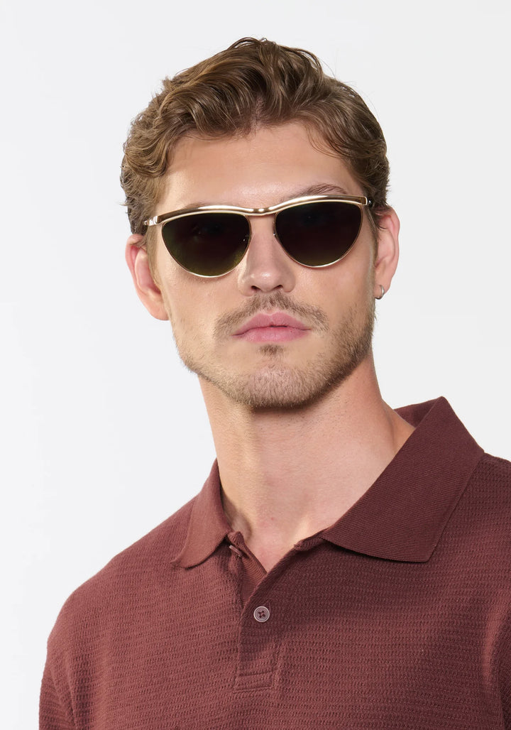 Man wearing gold-framed sunglasses and a brown polo shirt against a white background