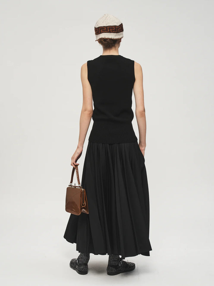 Person wearing a black sleeveless top and long black skirt with a brown bag on a white background
