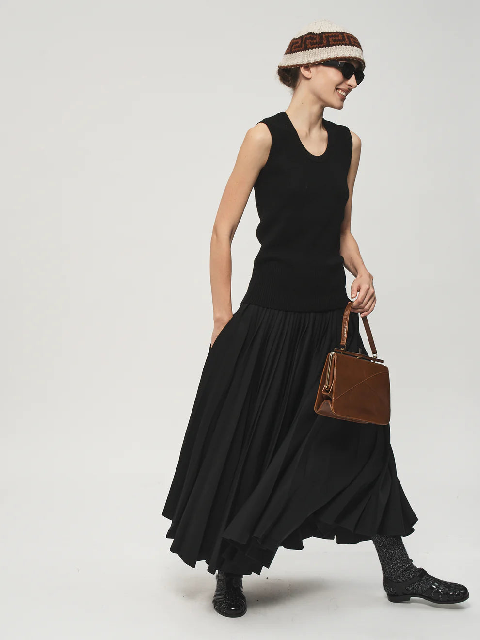 Woman wearing a black sleeveless top and long skirt with a brown bag on a light gray background
