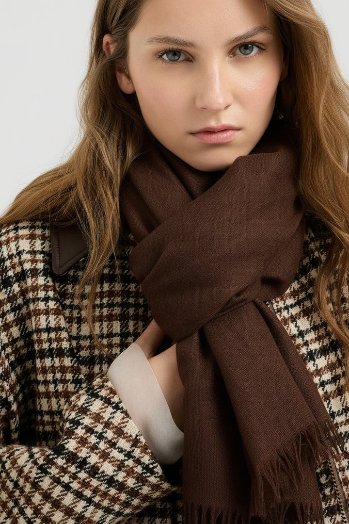 A close-up of a woman wearing a muted olive green scarf wrapped around her neck, paired with a brown, beige, and black plaid coat against a plain light gray background.