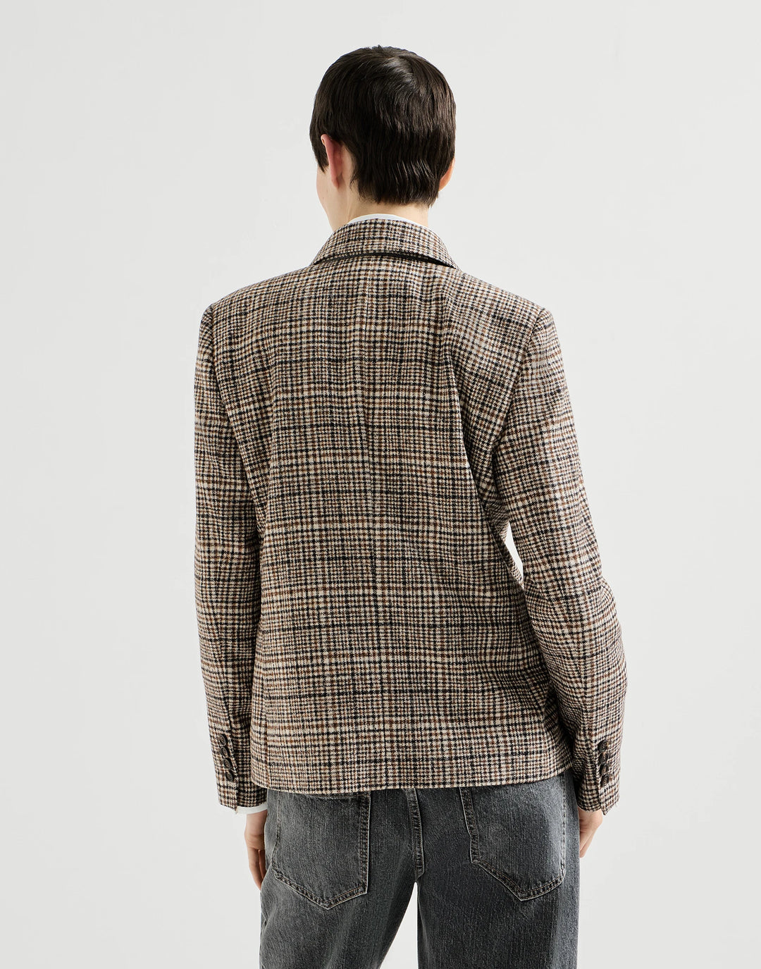 A man viewed from behind wearing a brown and beige plaid blazer with dark gray jeans, set against a plain light gray studio background.