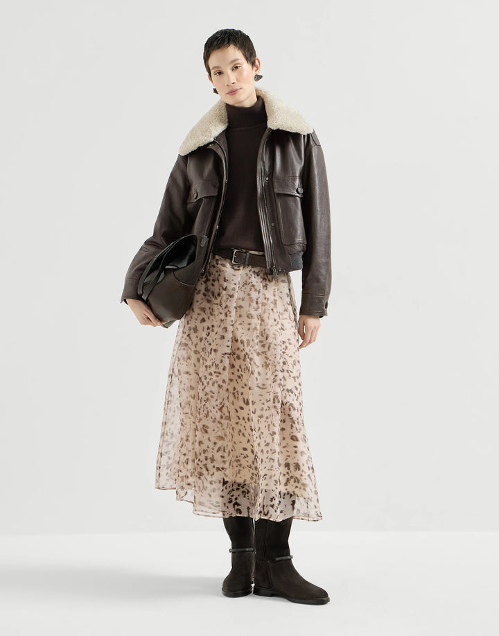 A model stands against a light gray studio background wearing a dark brown leather aviator-style jacket with a cream shearling collar, a black turtleneck, a beige leopard-print midi skirt, and black ankle boots.