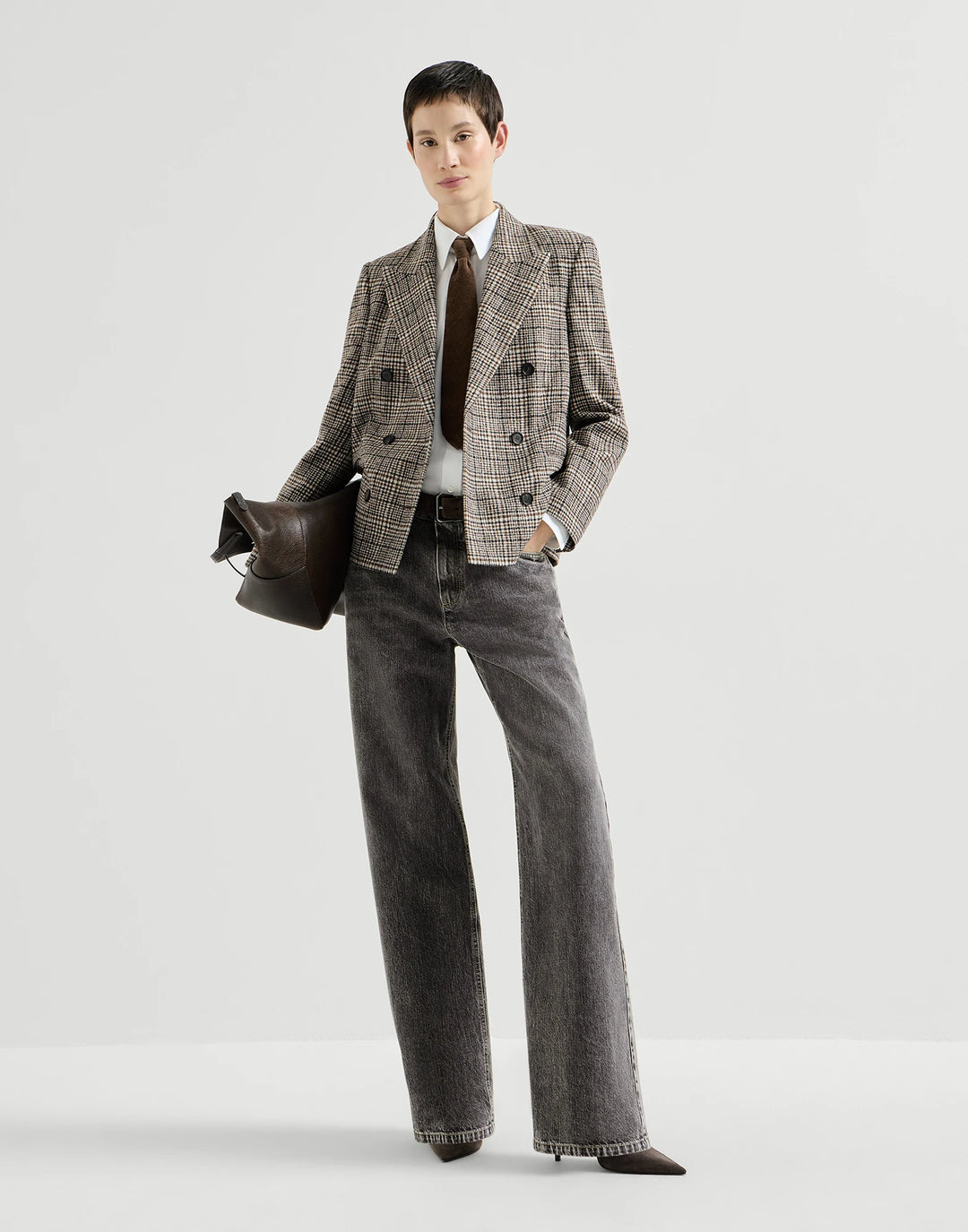 A model stands against a light gray studio background wearing a double-breasted plaid blazer in beige and brown tones, paired with dark gray wide-leg jeans, a white shirt, a dark brown tie, and carrying a dark brown leather bag.