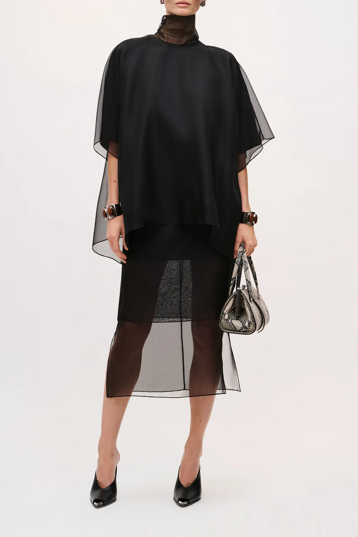 A model wearing a black sheer capelet overlay dress with a high mock neck, paired with sheer black tights and black pointed-toe heels, holding a metallic silver handbag against a plain white studio background.