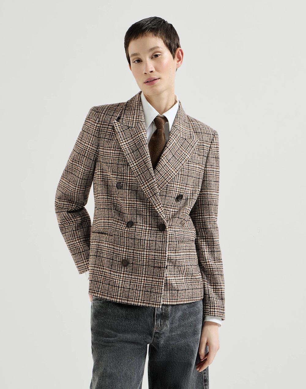 A model wearing a brown and beige plaid double-breasted blazer over a white shirt and brown tie, paired with dark gray jeans, against a light gray studio background.
