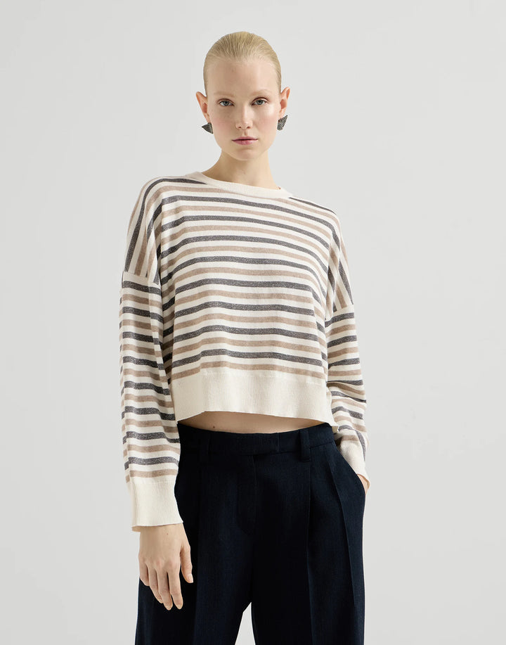 A model wearing a cropped, long-sleeve knit sweater with horizontal beige and brown stripes on an off-white base, paired with dark navy trousers, against a light gray studio background.