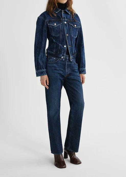 A model wearing a dark blue denim jacket and matching dark blue straight-leg jeans, paired with brown ankle boots, against a plain light gray studio background.