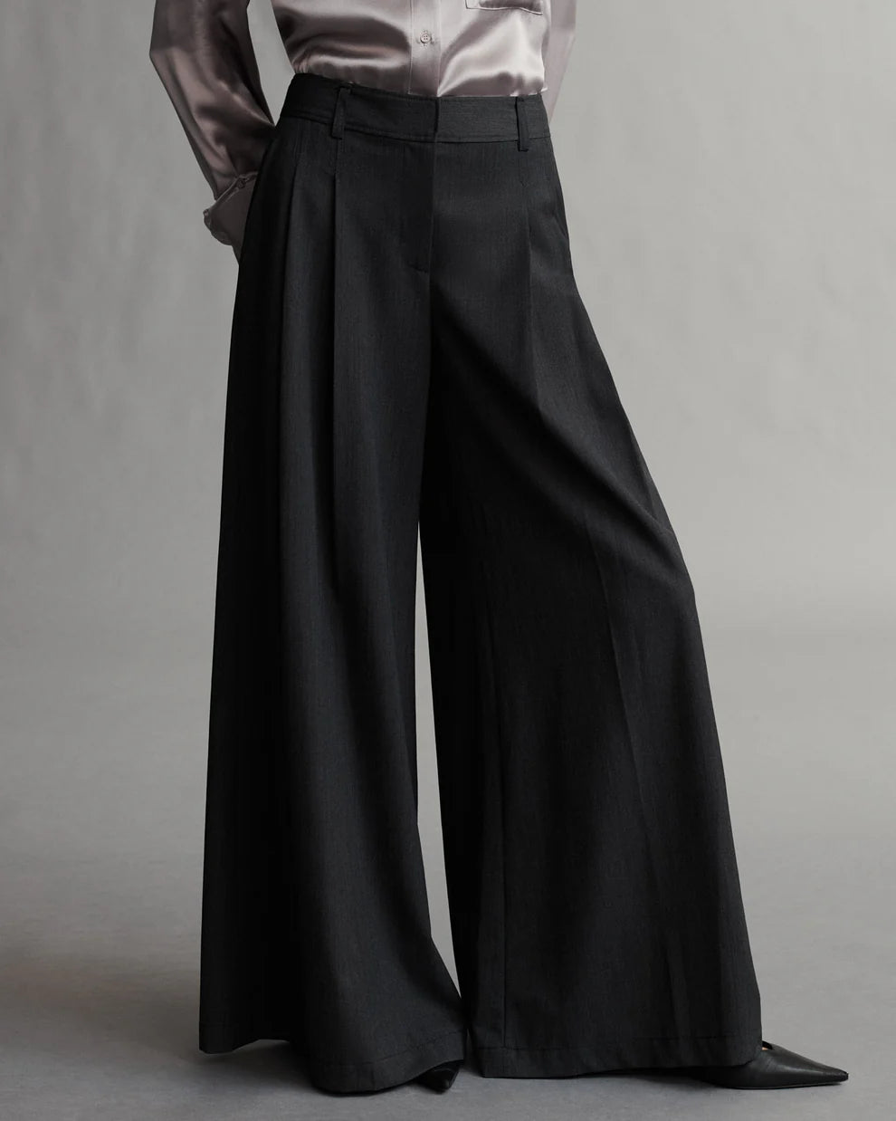 A model wearing black pleated pants with a wide leg silhouette, featuring side pockets and a zip-fly.