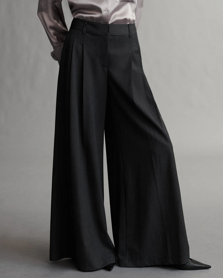 A model wearing black pleated pants with a wide leg silhouette, featuring side pockets and a zip-fly.