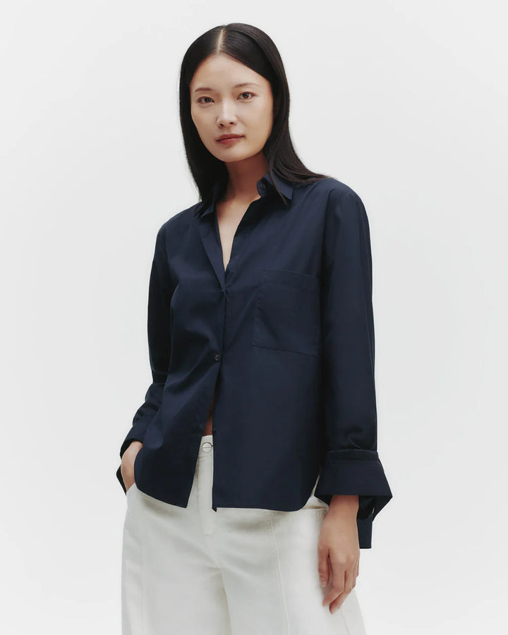 A woman modeling a navy oversized button down shirt with a center box pleat at the back, chest patch pocket, spread collar, and single roll button cuffs.