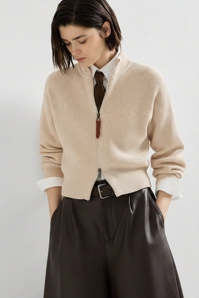 A woman wearing a ribbed knit zip-front cardigan in a light beige/cream color, styled over a white collared shirt, dark brown tie, and dark brown wide-leg trousers, against a plain light gray studio background.