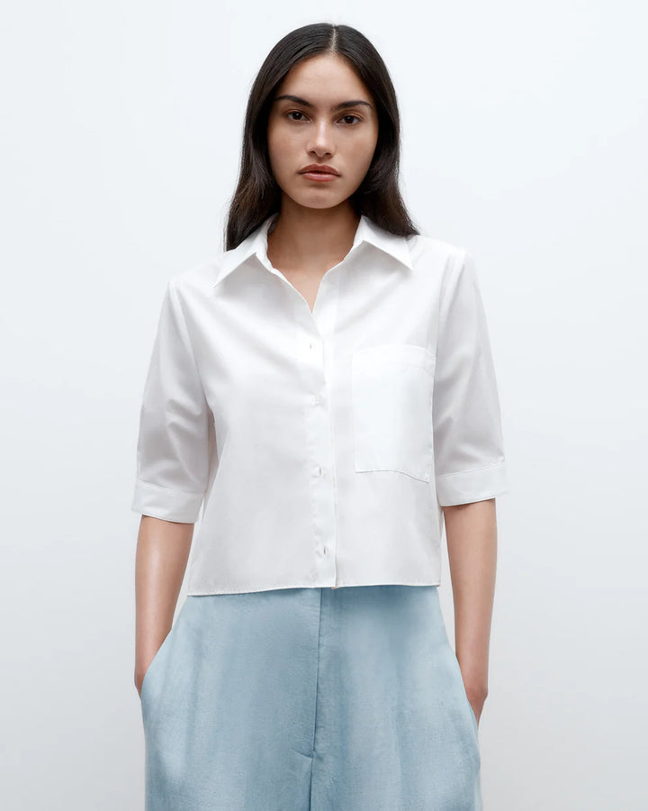 A woman wearing a white, short sleeve button-down shirt with a collar, chest patch pocket, and French cuffs. The shirt is cropped and made of cotton poplin with a smooth texture.