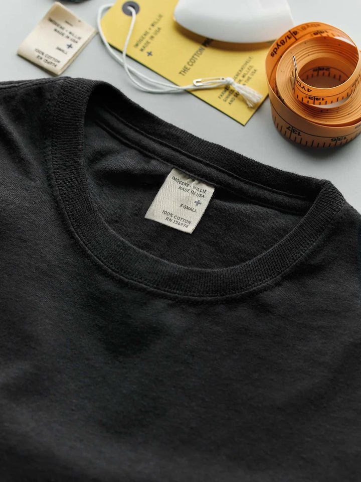 Black t-shirt with a tag on a gray surface, accompanied by a measuring tape and white object.