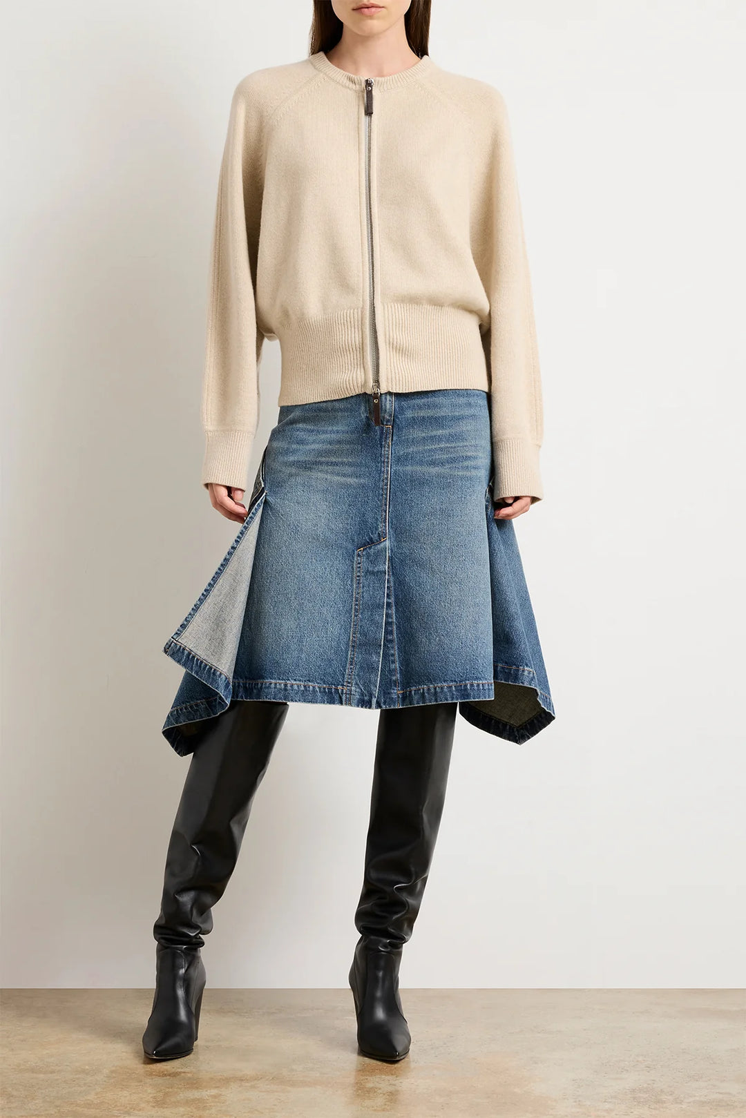Person wearing a beige zip-up sweater, blue denim skirt, and black pants on a plain background