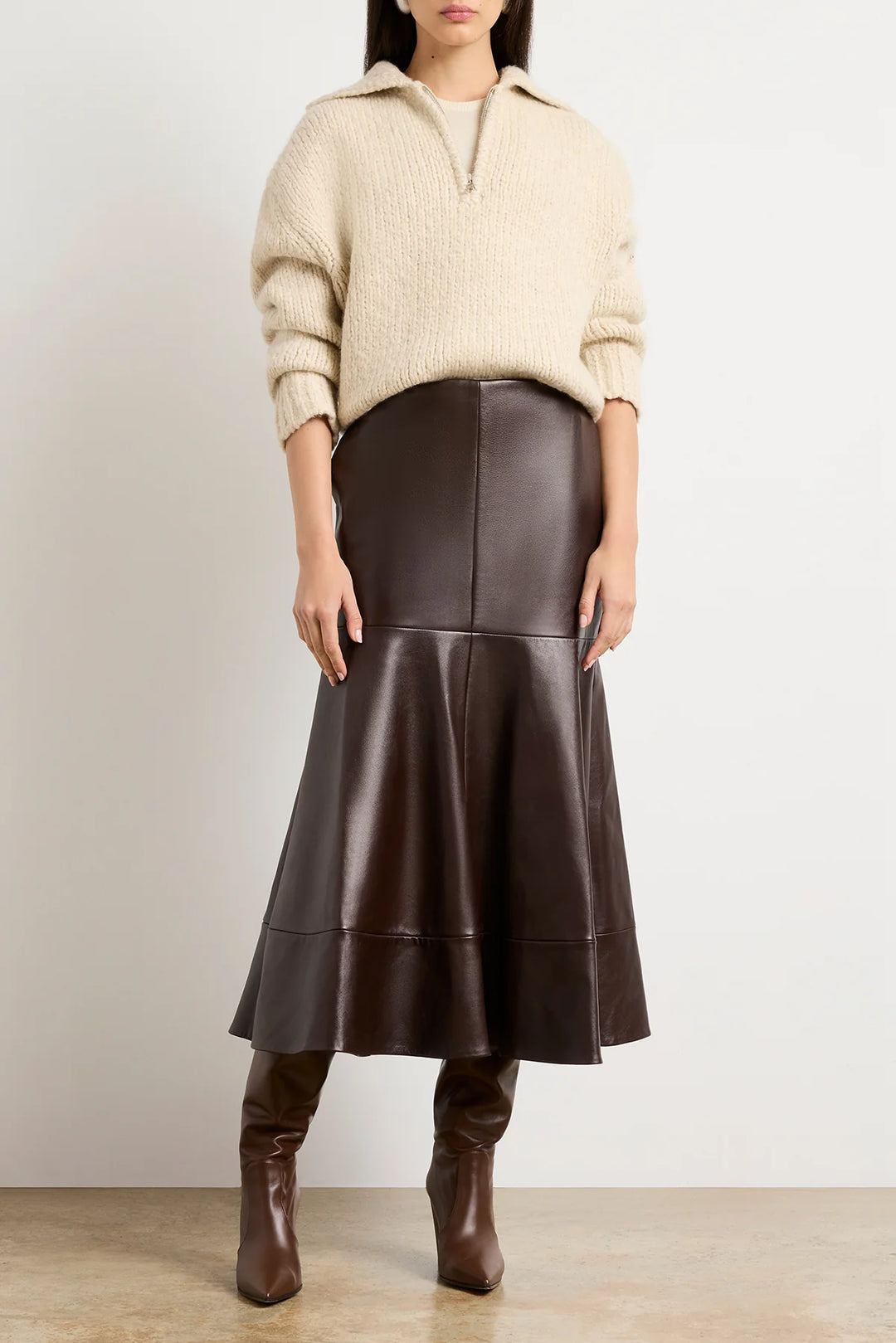 Person wearing a beige sweater and brown leather skirt with boots on a plain background