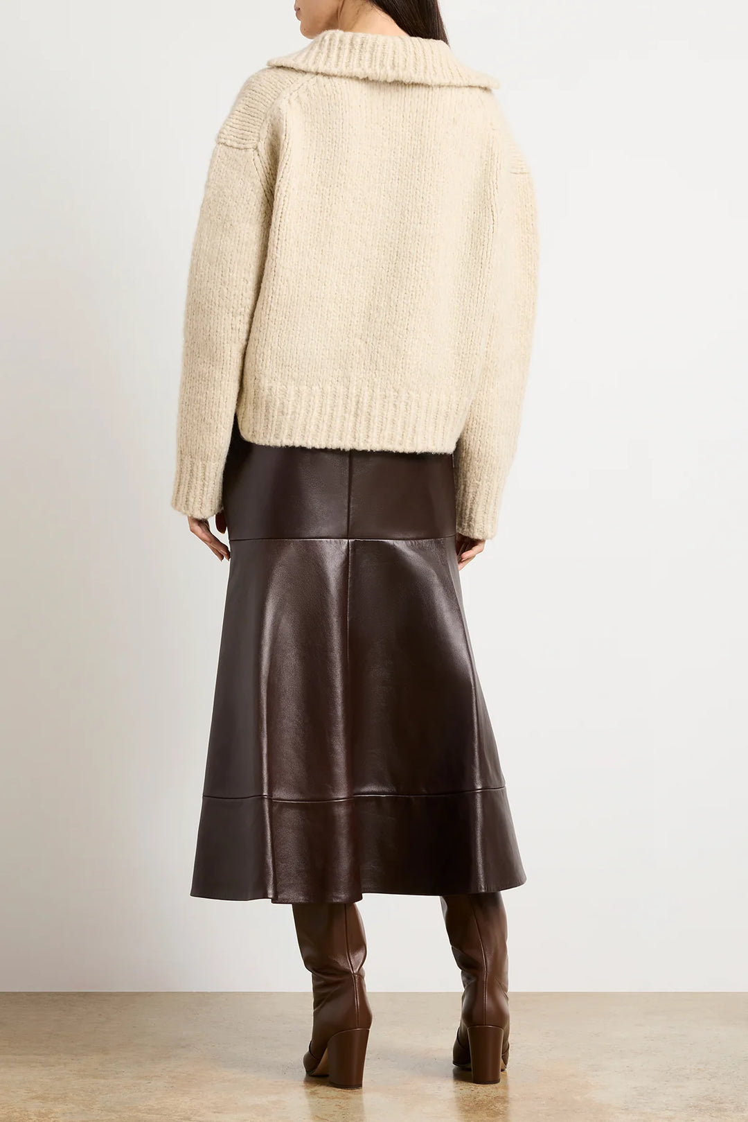 Person wearing a beige sweater and brown leather skirt with brown boots on a plain background