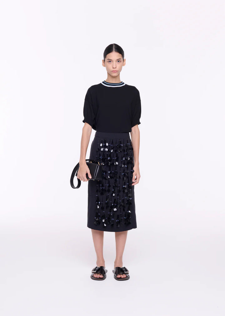 A model wearing a black midi skirt with sequin embroidery, paired with a black top and accessories.