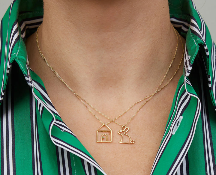 Gold necklace with house and rabbit pendant worn by a person in a green striped shirt.