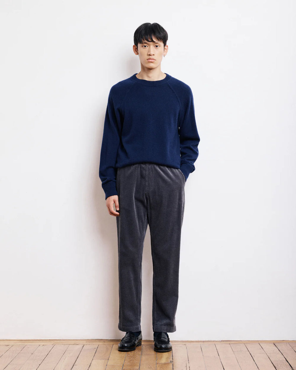 Person wearing a navy sweater and dark pants standing against a white wall.