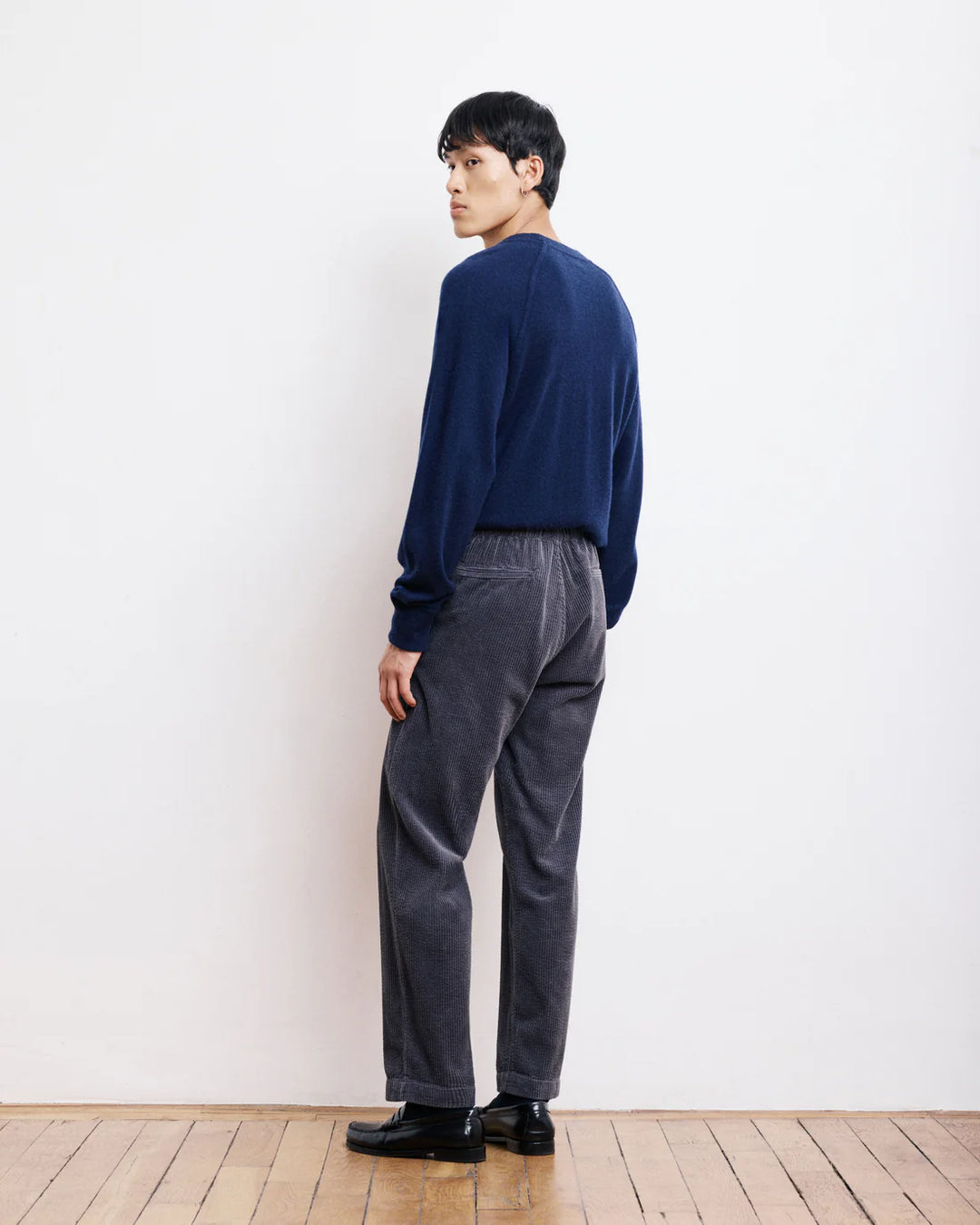 Person wearing a navy sweater and gray pants standing against a white wall.
