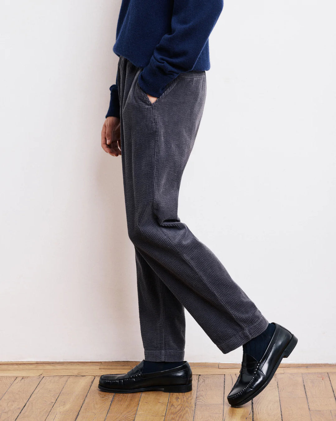 Person wearing dark gray pants and black shoes against a white wall.