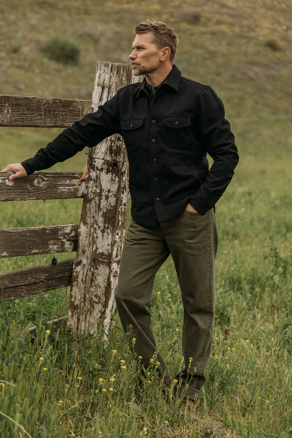 Man in a black jacket and green pants standing by a wooden fence in a grassy field.