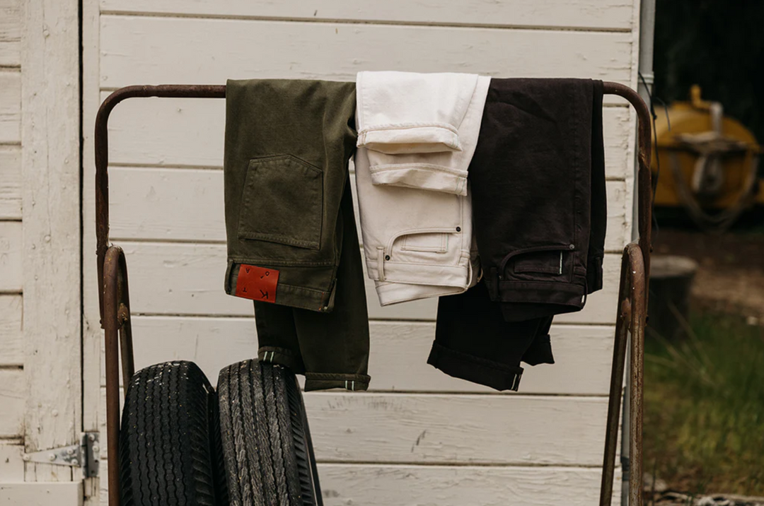 Three pairs of pants hanging on a metal rack against a wooden wall.