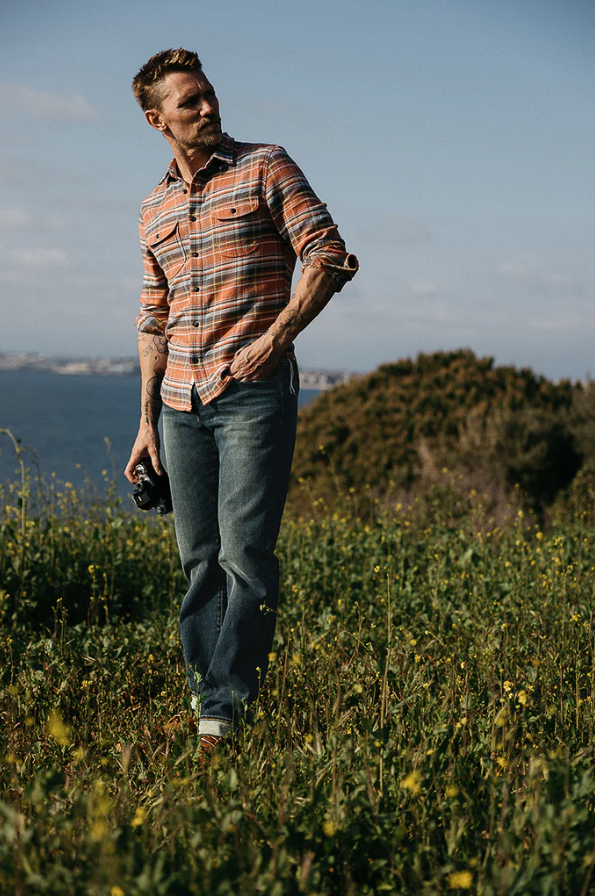 Man walking in a field wearing a plaid shirt and jeans, with a scenic background.
